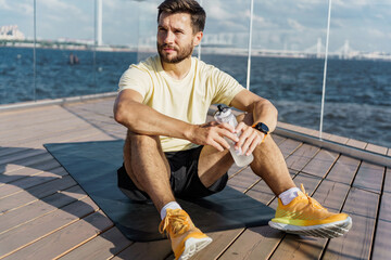 Young man sits on yoga mat by water, enjoying a refreshing break and hydration after workout