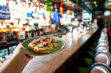  hand holds a plate of pizza in a vibrant bar setting, with a blurred background showcasing drinks and colorful lights, creating a lively atmosphere.
