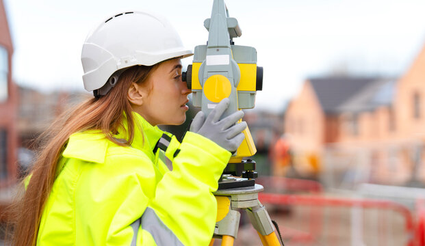 Portrait of young Woman in safety gear using a surveying instrument at a construction site while buildings are being developed nearby