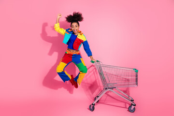 Young woman in colorful denim outfit enthusiastically jumping next to a shopping cart against a...