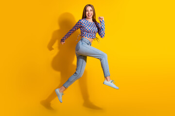 Cheerful young woman in casual jeans and colorful top jumping against yellow background with enthusiasm