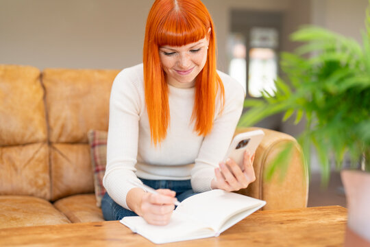 Woman with bright red hair writing in a notebook while sitting on a sofa with a plant in a cozy living room