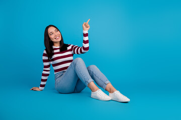 Smiling young woman pointing upward while sitting stylishly on a vibrant blue background.