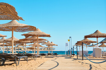 Beach umbrellas parasol made of wood and sunbeds standing on seashore against the backdrop of sunny blue sky and glare. View over tropical beach with rows of parasols. Vacation and Travel Concept.