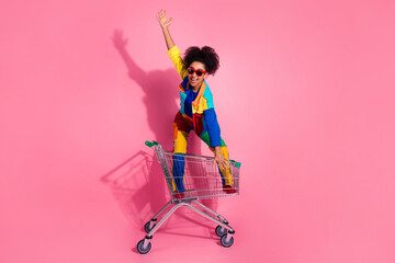 Young woman in colorful outfit excitedly posing with shopping cart on pink background