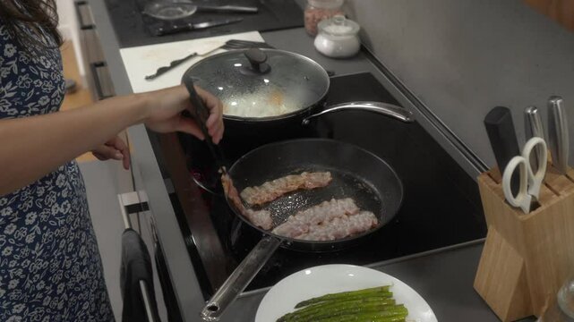 A woman is frying bacon on an induction stove in her kitchen. Using tongs, she flips sizzling strips of bacon in a hot non-stick pan. A bacon is cooking in its own fat, turning crispy and golden brown