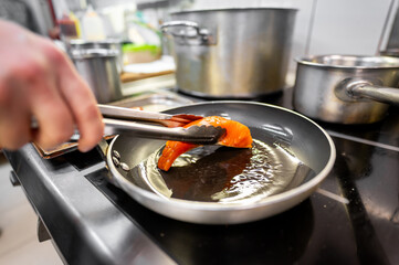 A close-up of a hand using tongs to place a piece of salmon into a heated frying pan on a modern kitchen stove, with pots in the background.