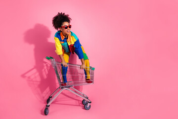 Stylish woman in vibrant clothing posing with shopping cart against a pink background