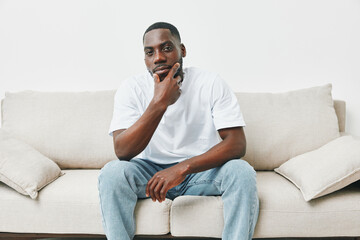 Thoughtful young man in casual white t shirt and jeans, sitting on a light colored sofa, reflecting on life and emotions in a minimalist setting