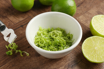 Lime zest in bowl, fresh fruits and zester tool on wooden table, closeup