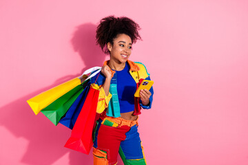 Stylish woman carrying colorful shopping bags while using her smartphone against a vibrant pink...