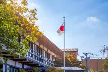 A Japanese flag waves atop a flagpole beside a traditional-style building, surrounded by lush greenery under a bright sky.
