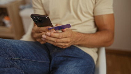 Young man shopping online with smartphone and credit card in modern apartment setting, focusing on digital transactions and casual lifestyle.