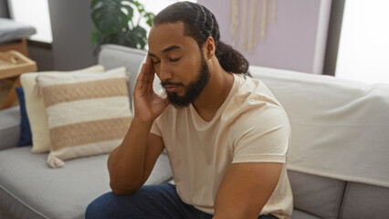 Young man with beard sitting in a living room looking thoughtful, surrounded by soft furnishings,...
