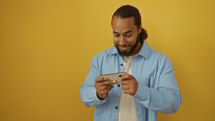 Man smiling at smartphone over yellow background, wearing blue shirt, beard visible, playing game or watching video, isolated in vibrant setting, adult enjoying digital content.