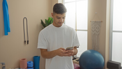 Young man in gym checking phone near exercise equipment and yoga balls, conveying fitness, technology use, and indoor activity in an athletic setting.
