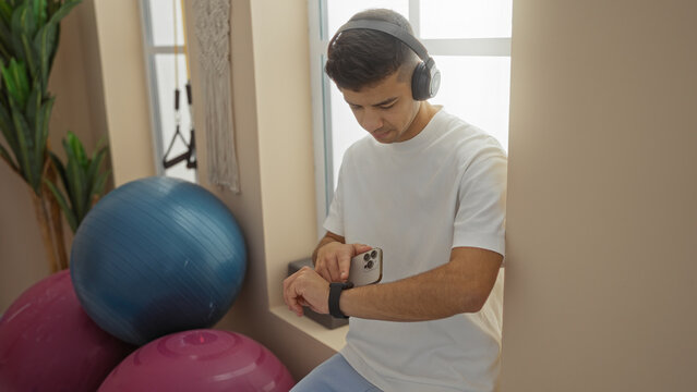 Young man wearing headphones checks smartwatch in a bright gym surrounded by exercise balls and plants, capturing a relaxed fitness moment.