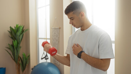 Young man exercising with dumbbell in gymnasium surrounded by fitness equipment and plants in a...