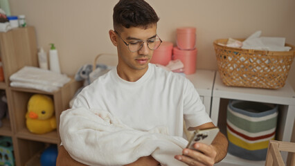 Young man in glasses cradles newborn in cozy room while checking phone, surrounded by baby supplies in a peaceful, nurturing atmosphere.