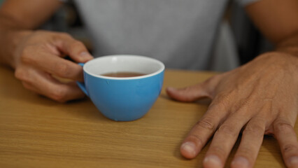 Young hispanic man holding blue cup in office setting with focus on hands resting on wooden table indicating a casual professional environment indoors.