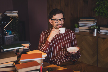 Young man enjoying a warm beverage while surrounded by books in a cozy home office interior with a casual and stylish ambiance.