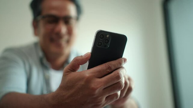 Close up asian senior middle aged man holding smartphone while sitting on sofa in living room at home, elderly man with mobile phone for social web application online. 

