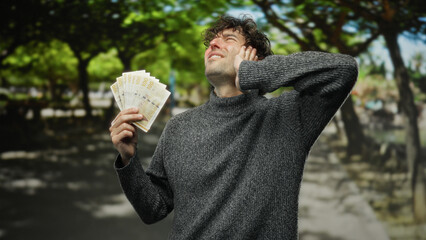 Hispanic man in park holding norwegian krone appears pained while outdoors surrounded by nature in...