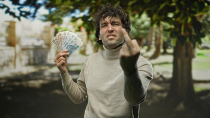 Hispanic man outdoors in a park holding polish zloty banknotes with an angry expression,...