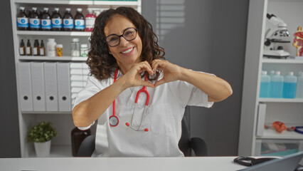 Hispanic woman doctor in a hospital room forming a heart shape with her hands, displaying warmth and professionalism surrounded by medical equipment and supplies.