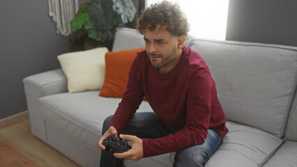 Young man playing video games on a gray sofa in a cozy living room, surrounded by colorful...