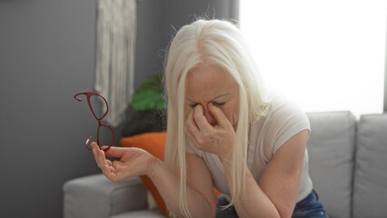 Woman with albinism holding glasses and rubbing eyes in a cozy living room, suggesting fatigue and...