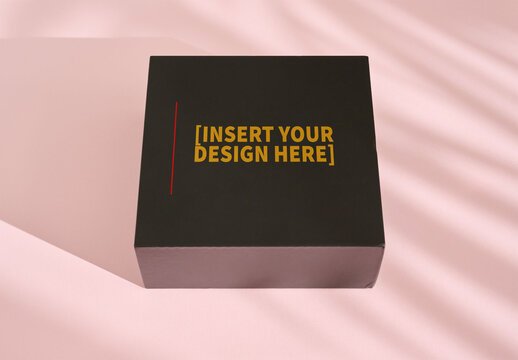 A black square box with a shiny texture rests on a pale surface, creating soft shadows in warm light.