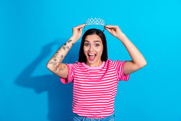 Smiling young woman in casual pink striped shirt posing with a tiara against a blue background,...