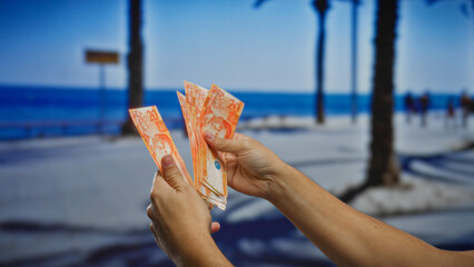 Hands holding philippine banknotes outdoors at a seaside location, with palm trees and a beach in the background, suggesting a vibrant day in a coastal city.