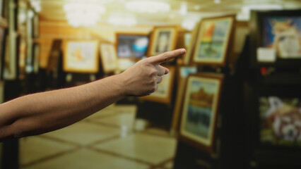Man's hand pointing at artwork in a brightly lit art gallery room, showcasing vivid paintings displayed in an elegant museum setting.
