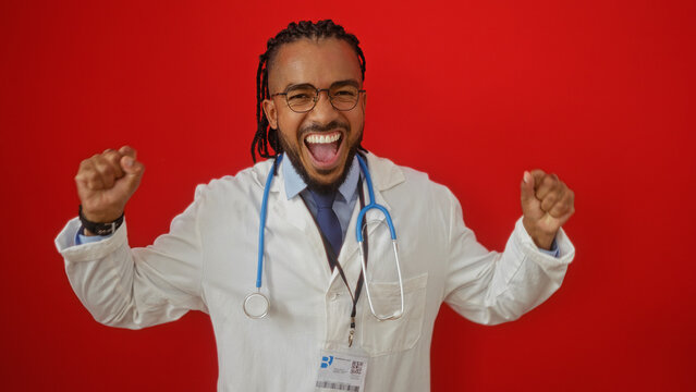 Young man in white coat with braids and stethoscope smiling, expressing joy against isolated red background, highlighting professional and confident demeanor.