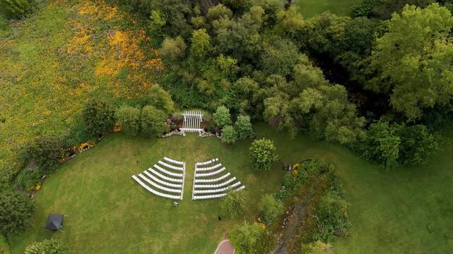 The aerial drone view of the outdoor wedding setup with rows of white chairs facing a wooden arch. The setting is on a grassy area, surrounded by trees, under a clear blue sky. Wide view