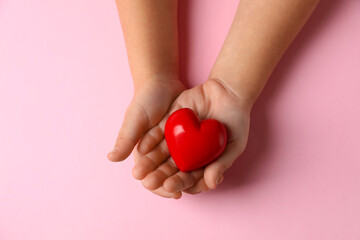 Child holding decorative red heart on light pink background, top view