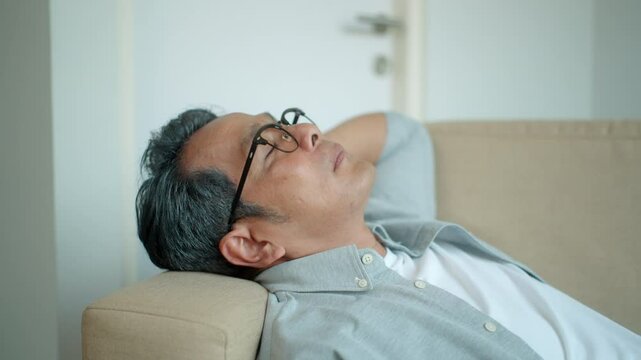 Asian senior middle aged man resting and eye closed on comfy sofa in living room at home, calm old mature male relaxing napping daytime with no stressed on couch at weekend.
