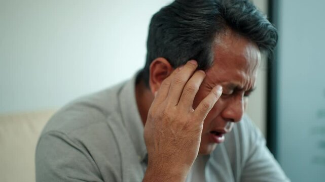 Asian senior mature man suffering from headache while sitting on sofa in living room at home, elderly male feeling pain with migraine, health care and medical insurance for retirement people concept.

