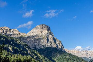 Obraz premium The striking Sassongher peak in the Dolomites towers over dense green forests, framed by a vivid blue sky and steep rocky terrain