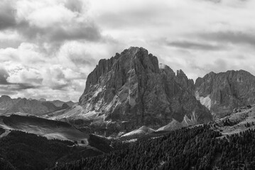 A dramatic black and white view of Sassolungo in Val Gardena, showcasing its rugged cliffs, dense forests, and moody clouds for a timeless alpine scene
