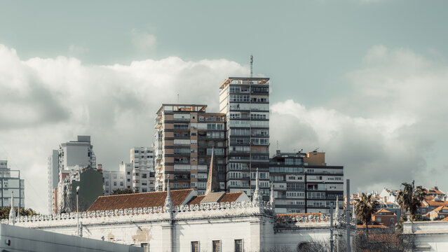 Modern high-rise residential buildings with balconies and glass windows stand against a cloudy sky, contrasting with historic white architecture featuring ornate details and terracotta roofs