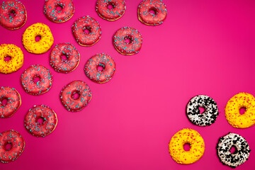 A flat lay of colorful donuts scattered on a vibrant pink background. The mix of pink, yellow, and cookies-and-cream toppings creates a modern, stylish, and appetizing food composition.