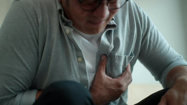 Close up senior mature man touching his chest from suffering heart attack while sitting alone on sofa in living room at home, old male having cardiac disease problem, medical insurance for retirement.