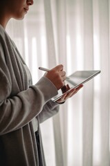Latina woman using a tablet and stylus, surrounded by soft, natural lighting and sheer curtains.