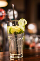 A close-up shot of a classic mojito served in a tall glass, filled with lime slices, fresh mint, and ice cubes, with a black straw.The background is blurred, highlighting the drink’s freshness. 