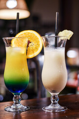 A close-up of two tropical cocktails on a bar counter. One is a layered, multi-colored drink with blue, green, and yellow hues, garnished with an orange slice.  Piña Colada with a pineapple wedge.
