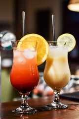 A close-up of two tropical cocktails on a bar counter. Tequila Sunrise with orange slice and Piña Colada with a pineapple wedge. Bokeh background in bar.