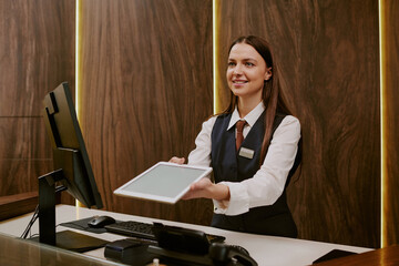 Medium close up of smiling young female receptionist in smart clothes standing behind reception...